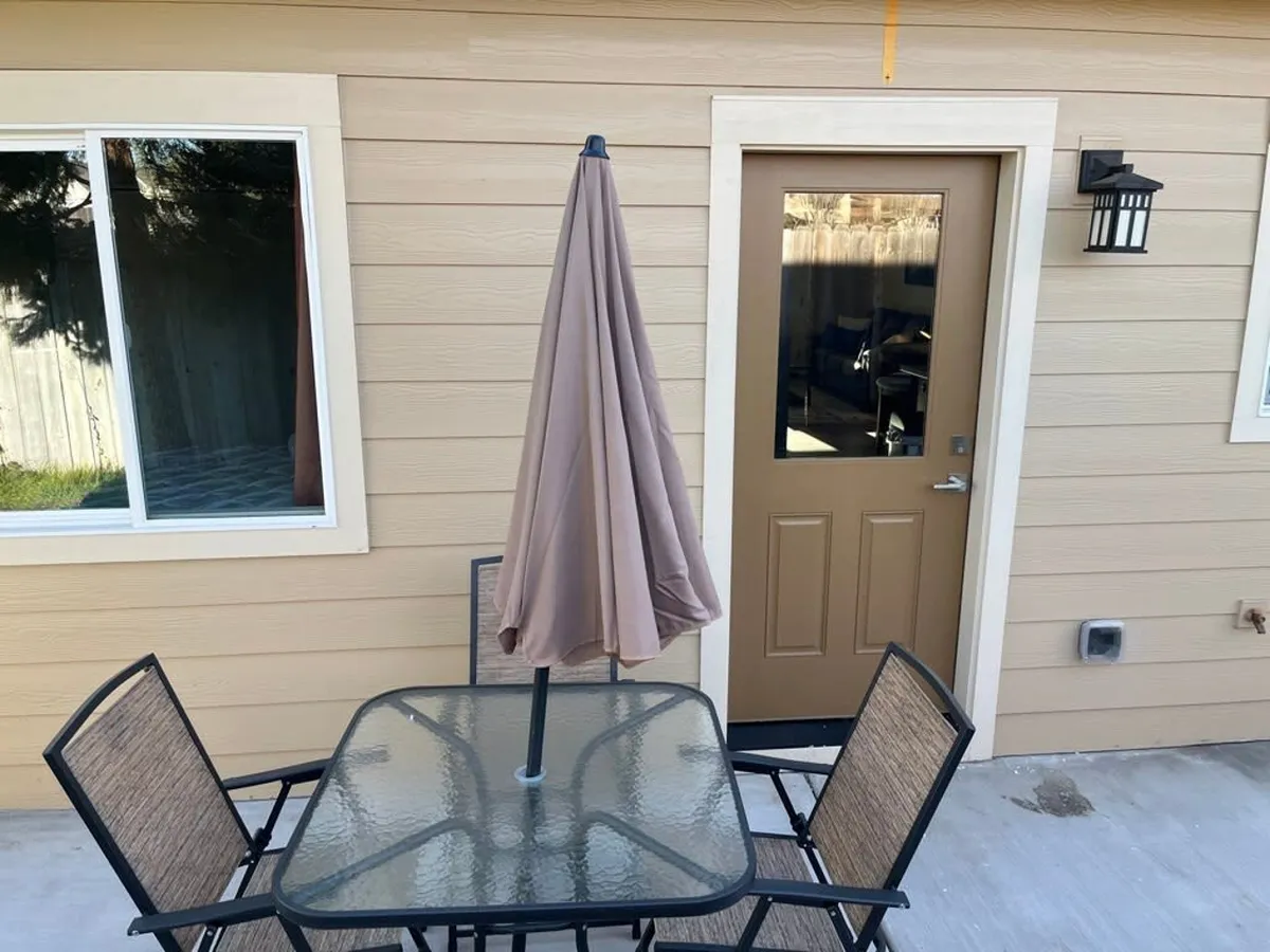 An exterior view shows a brown door leading into the cottage, flanked by a window, with a glass-top patio table and umbrella nearby. The area is suitable for parking, and the unfinished yard is visible, adding transparency to the home's current status.