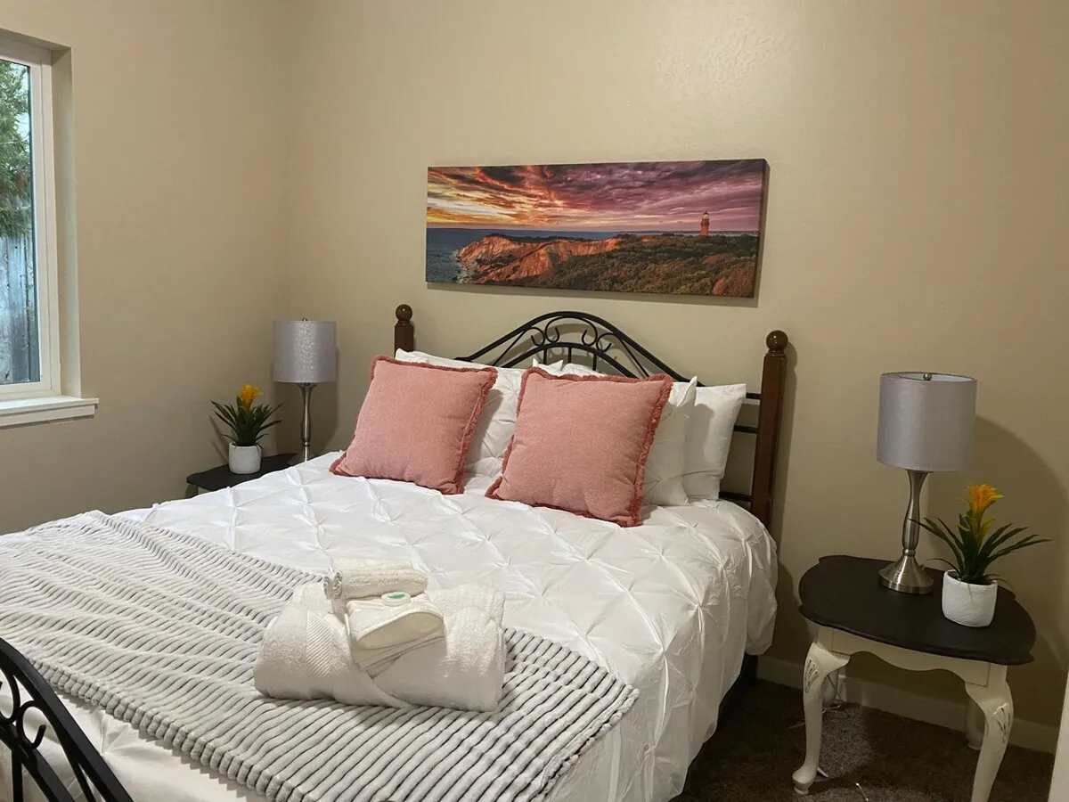 A cozy bedroom with a queen bed dressed in crisp linens and decorative pillows, complemented by a landscape artwork above the headboard and a nightstand with a lamp. The overall vibe is welcoming and serene, with natural light enhancing the tranquil ambiance.