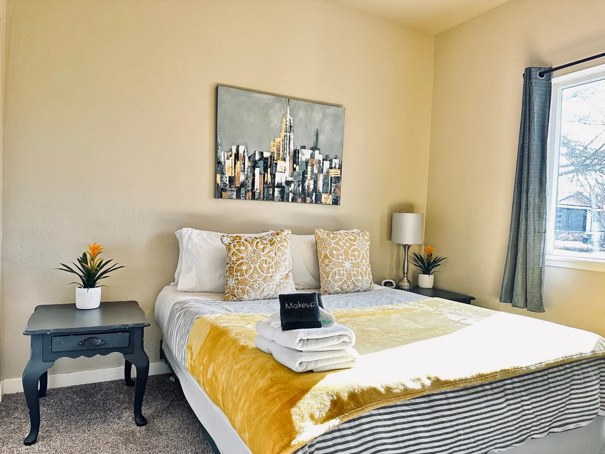 A serene bedroom featuring a queen bed adorned with decorative pillows, a cityscape painting above the headboard, a decorative lamp on a nightstand, and a small potted plant. The space is bathed in soft natural light, evoking a tranquil and restful atmosphere.