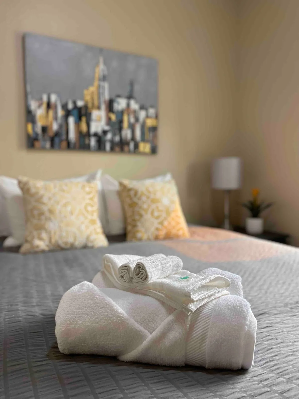A neatly arranged stack of fluffy towels sits atop a shelf, accented by stylish decorative pillows and a striking cityscape painting. The textures and organization suggest both comfort and attention to detail, indicative of well-thought-out storage and the presence of laundry amenities.