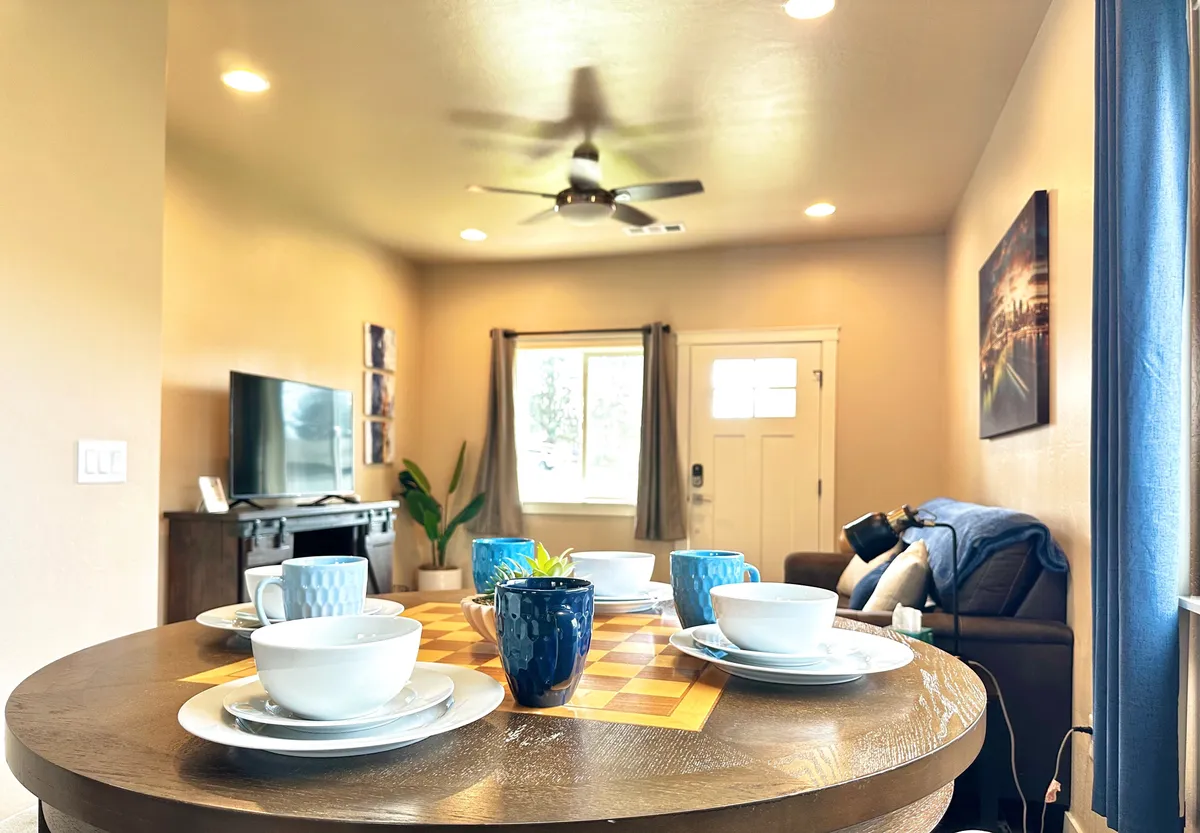 This scene presents an inviting open-concept space featuring a dining table set for meals, a comfortable couch with a throw blanket, a television, and a large window draped with curtains. The setup is both functional and aesthetically pleasing, designed for gatherings, family meals, or unwinding after a day of adventure. The natural light streaming in underscores the fresh, brand-new atmosphere.