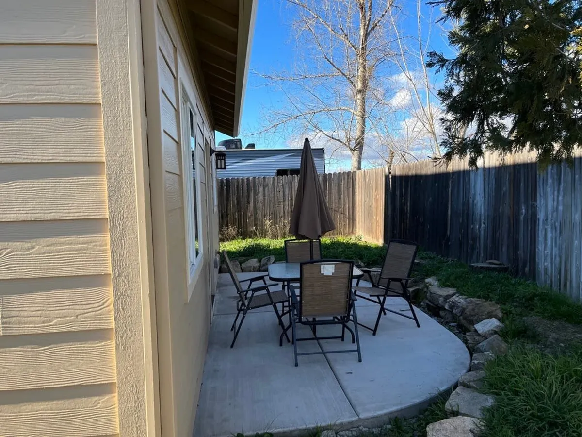 This image depicts a welcoming outdoor patio area featuring a table with an umbrella, several chairs, a fenced yard, and mature trees in the background. The setting suggests opportunities for outdoor dining, relaxing, and casual gatherings, all within the privacy of the home’s yard.