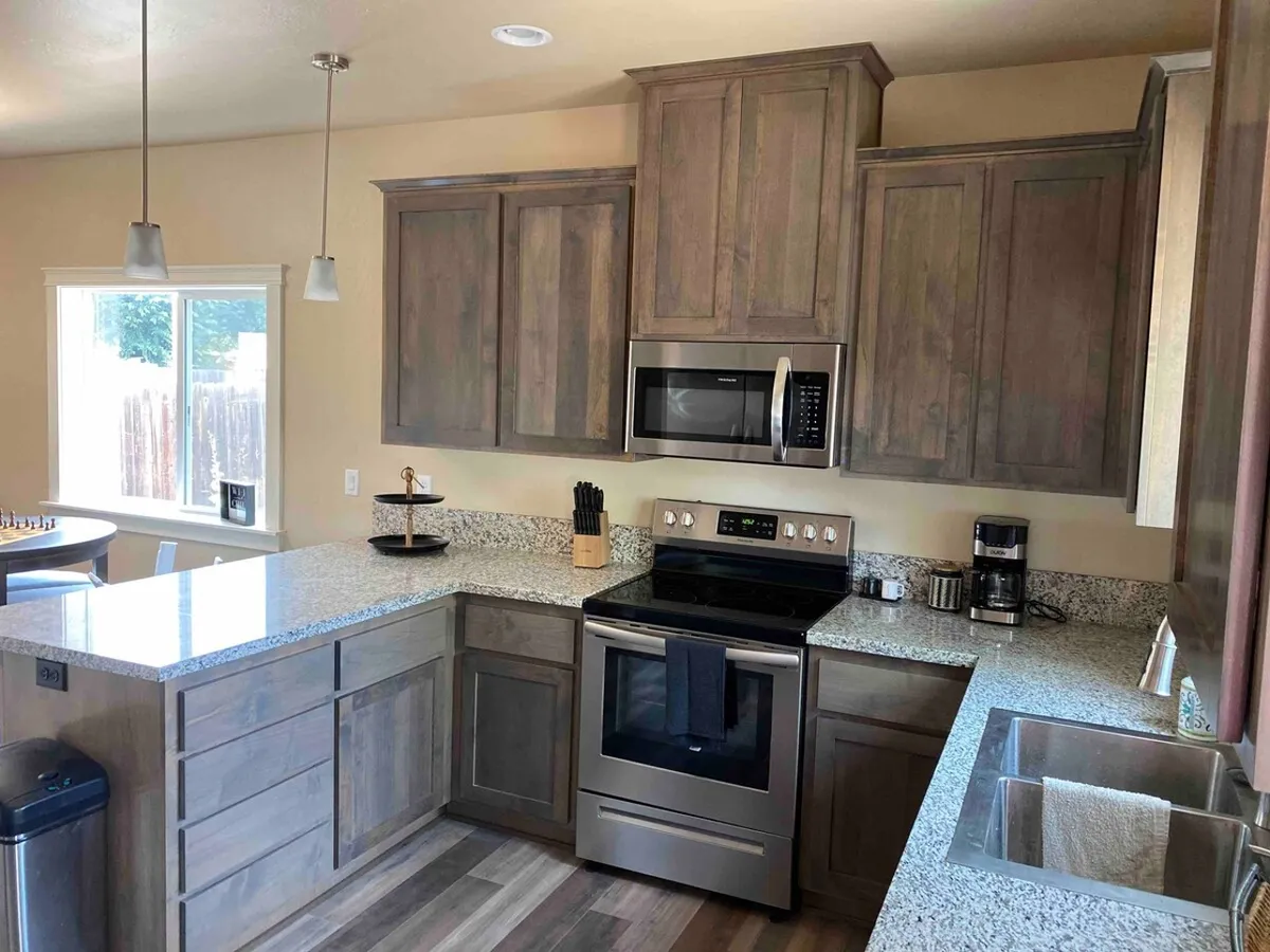 A spacious, well-lit kitchen featuring granite countertops, stainless steel appliances, wooden cabinets, and a window letting in natural light. The space feels warm and is clearly set up for cooking and gathering, with ample room for meal preparation and casual dining.