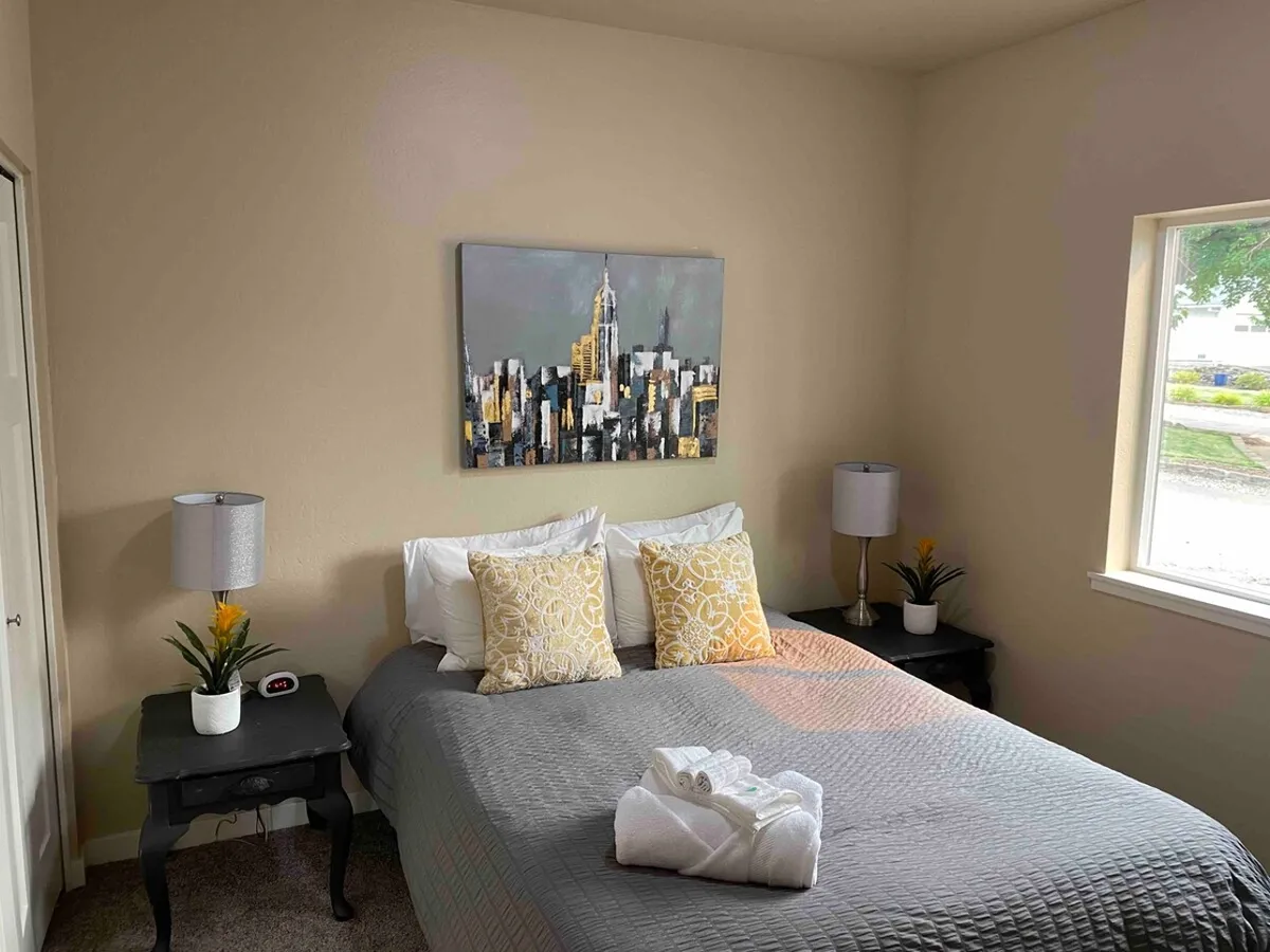 A sunlit bedroom features a bed with decorative pillows, a large window letting in natural light, and a cityscape painting. The bed is adjacent to what could be built-in storage solutions, reflecting a welcoming and organized atmosphere.
