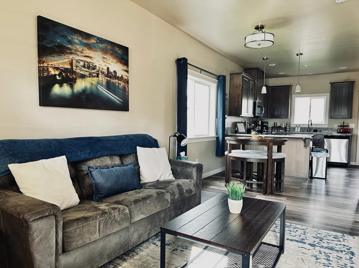 The image showcases a bright, inviting living space with a plush sofa, stylish coffee table, tasteful wall art, and an open kitchen area. The room exudes comfort and warmth, making it ideal for relaxing, small gatherings, or quiet evenings at home.