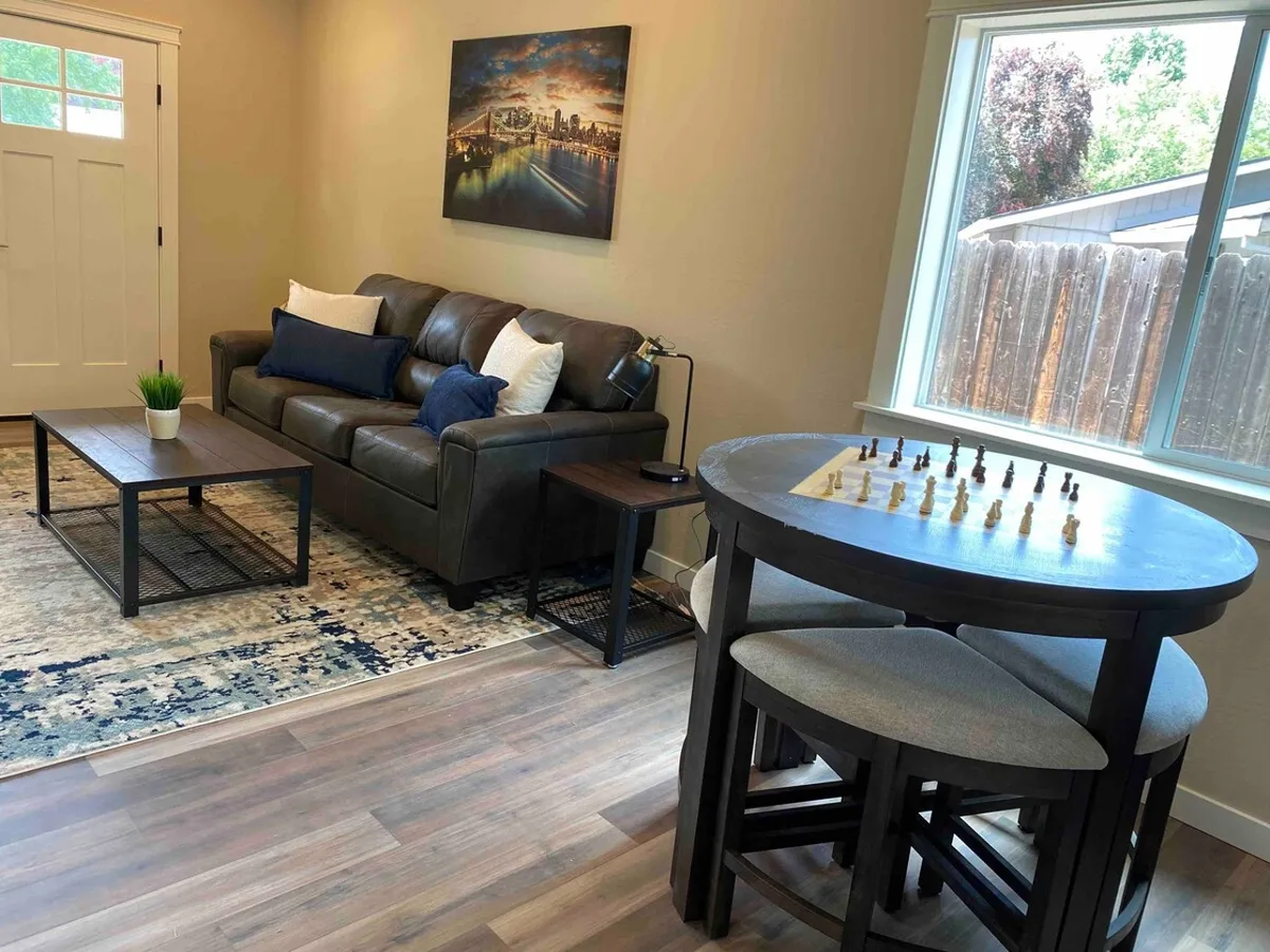A cozy living area featuring a comfortable sofa, a chess table set for a game, eye-catching artwork on the walls, and a coffee table. The arrangement encourages relaxed socializing or quiet downtime, with natural light hinting at a welcoming, homey atmosphere.